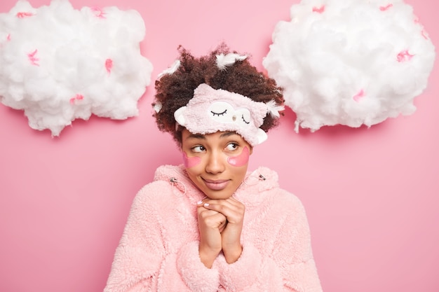 portrait of thoughtful woman in sleepwear keeps hands under chin looks with pensive expression aside wears sleepmask undergoes skin care procedures applies beauty pads after sleeping