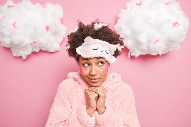 Portrait of thoughtful woman in sleepwear keeps hands under chin looks with pensive expression aside wears sleepmask undergoes skin care procedures applies beauty pads after sleeping