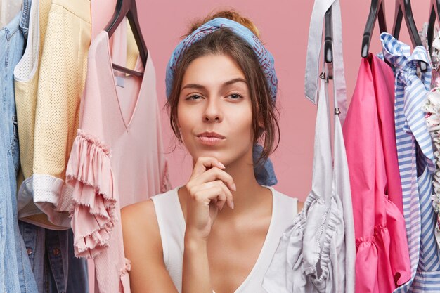 Portrait of thoughtful woman with scarf on head, standing near hangers with clothes, thinking over what to buy. Female shopaholic in boutique with pensive expression, having difficult choice