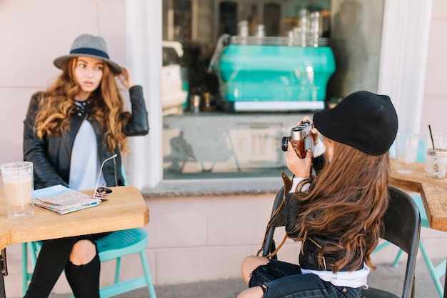 彼女の娘が彼女の写真を撮っている間コーヒーを飲みながらテーブルに座っているフェルト帽子で思いやりのある若い女性の肖像画。
