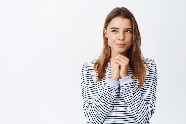 Portrait of thoughtful young woman with blond hair, squinting pensive, having interesting idea, looking left at copy space, standing over white wall