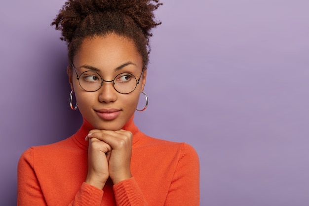 Portrait of thougthful Afro female keeps hands together under chin, wears round spectacles, earrings and orange turtleneck,  isolated over purple background