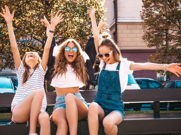 Portrait of three young beautiful smiling hipster girls in trendy summer clothes.Sexy carefree women sitting on the bench in the street.Positive models having fun in sunglasses.Raising hands