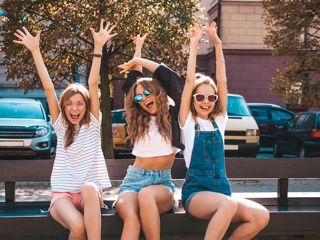 Portrait of three young beautiful smiling hipster girls in trendy summer clothes.Sexy carefree women sitting on the bench in the street.Positive models having fun in sunglasses.Raising hands