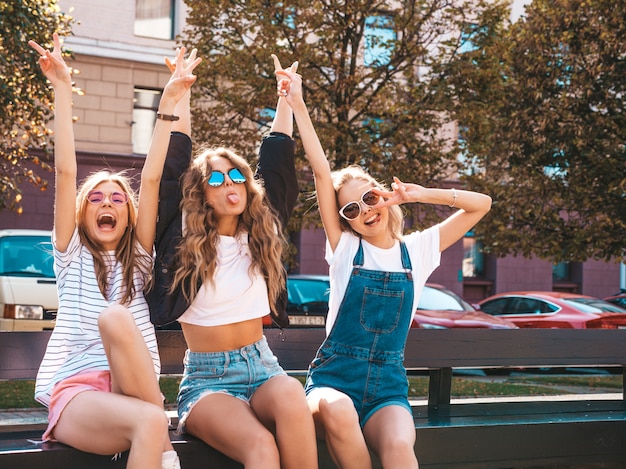 Портрет трех молодых красивых улыбающихся хипстерских девочек в модной летней одежде. Сексуальные беззаботные женщины, сидящие на скамье на улице. Позитивные модели, весело проводящие время в солнцезащитных очках. Поднимая руки