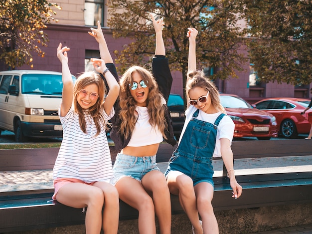 Portrait of three young beautiful smiling hipster girls in trendy summer clothes.Sexy carefree women sitting on the bench in the street.Positive models having fun in sunglasses.Raising hands