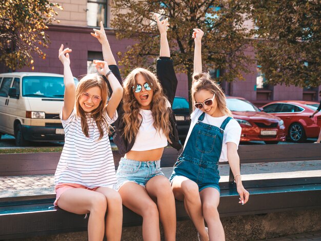 Портрет трех молодых красивых улыбающихся хипстерских девочек в модной летней одежде. Сексуальные беззаботные женщины, сидящие на скамье на улице. Позитивные модели, весело проводящие время в солнцезащитных очках. Поднимая руки