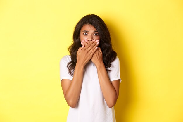 Portrait of timid and scared african-american girl, shut mouth and looking anxious, standing frightened over yellow background.