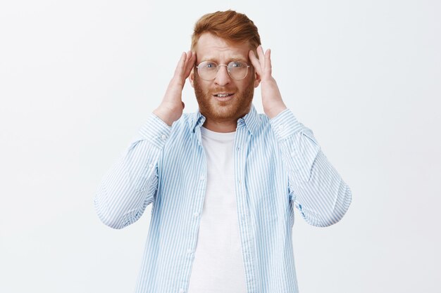 Portrait of tired good-looking male entrepreneur with ginger hair and beard in glasses, holding hands on temples and staring unfocused, having headache