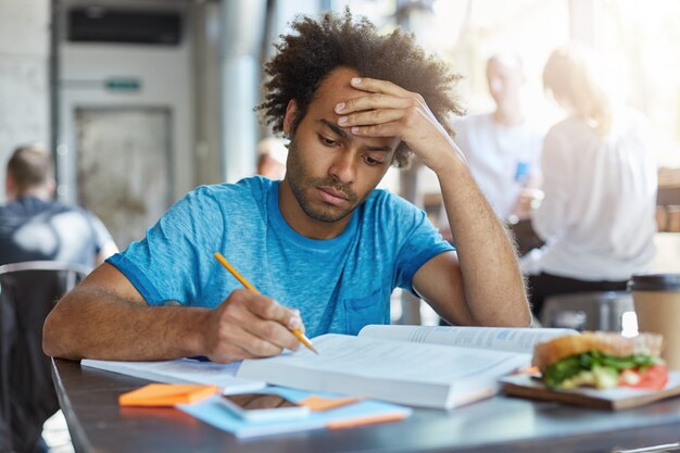 Portrait of tired serious mixed race male dressed casually sitting at wooden desk in cafe writing notes and reading books having lunch eating hamburger holding hand on head trying to concentrate