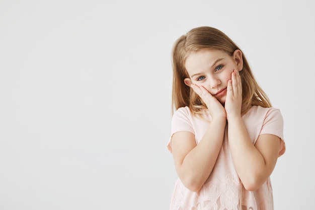 Portrait of tired sleepy little blond girl with blue eyes in pink beautiful dress, holding face with hands and looking . Copy space.