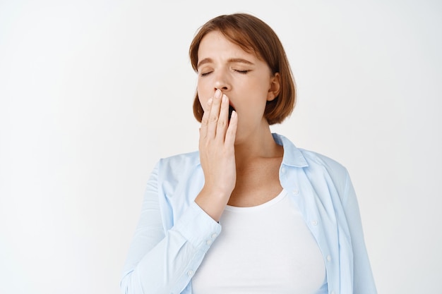 Portrait of tired young girl yawning, covering opened mouth with hand and close eyes, having fatigue after work, standing on white wall