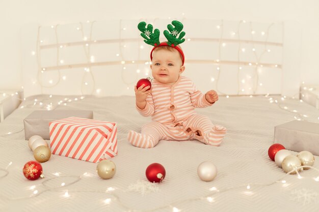 Portrait of toddler girl sitting on decorated bed and wearing santa striped long sleeve baby sleeper and festive deer horns, holding Christmas ball for decorations.