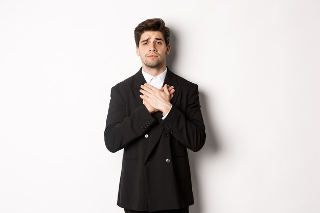Portrait of touched and compassionate guy in suit, holding hands on heart and looking with pity at camera, standing over white background.