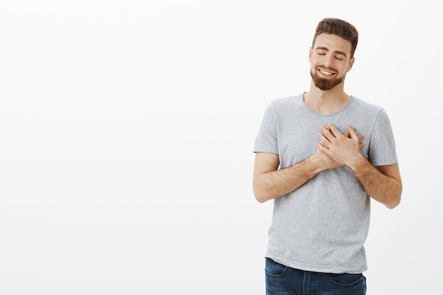 Ritratto di fidanzato commosso e felice con barba e acconciatura moderna, chiudendo gli occhi e sorridendo tenendo le palme sul cuore sognando ad occhi aperti essendo nostalgico e felice ricordando bei ricordi d'amore