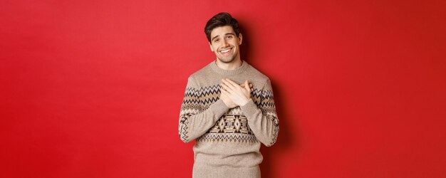 Portrait of touched and happy handsome guy receiving new year gift holding hands on heart and smiling saying thank you standing in christmas sweater against red background