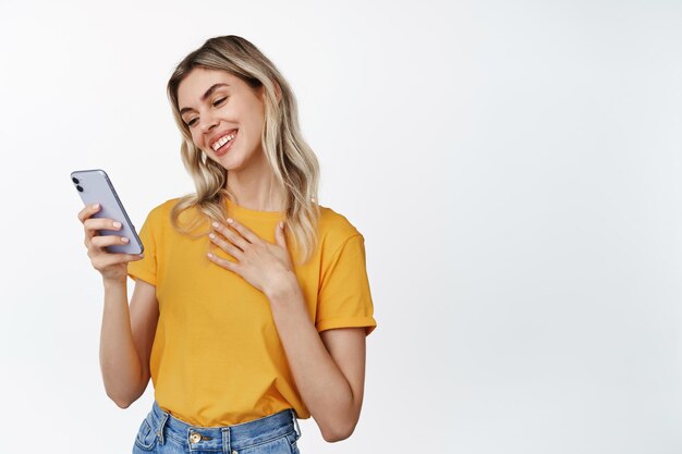 Portrait of touched young woman fond of something on smartphone screen reading message on mobile phone with delighted smile white background