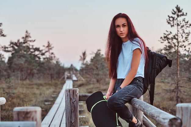Portrait of a tourist girl in a white shirt and jeans leaning on a wooden fence in a beautiful autumn meadow at sunset.