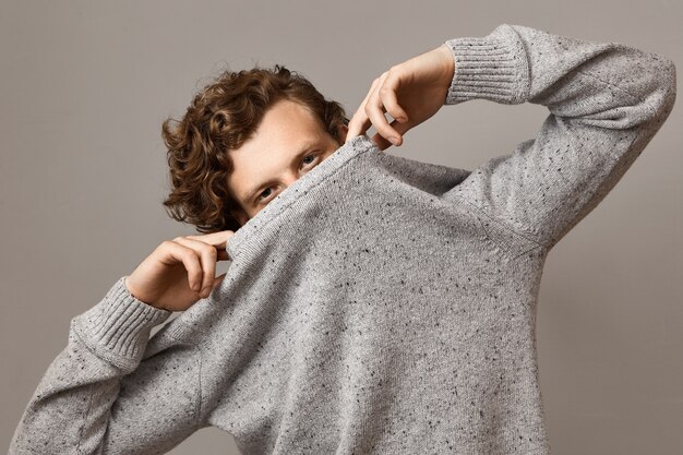 Free photo portrait of trendy looking cute young caucasian male with wavy voluminous curly hairstyle fooling around, pulling gray sweater over his face, leaving eyes open, having confident facial expression