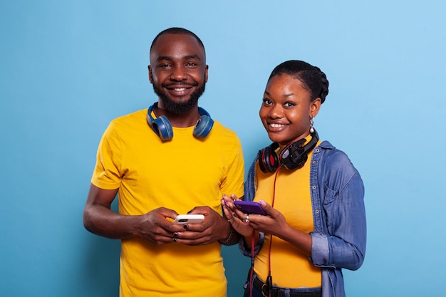 Portrait of trendy people holding mobile phone in hand and smiling at camera. Couple of millennials enjoying social media on smartphone and ignoring each other. Modern relationship