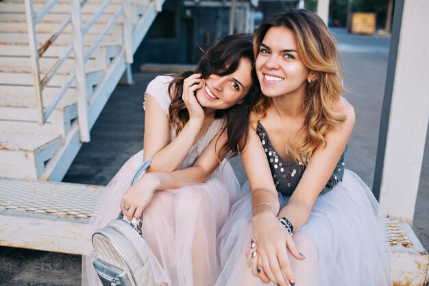 Portrait of two attractive girls in tulle skirts sitting outdoor on stairs. They smiling .