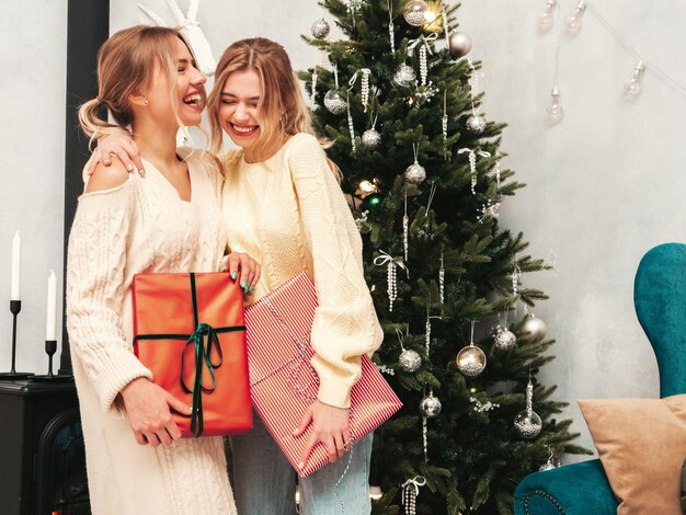2人の美しい金髪の女性の肖像新年の前夜に飾られたクリスマスツリーの近くでポーズをとるモデル暖かい冬のセーターを着た友人がお祝いの準備を楽しんでいます