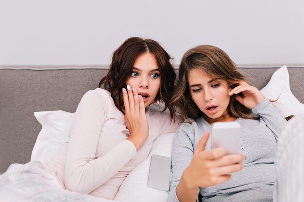 Portrait two beautiful girls in pajamas lying on bed. They looking on phone are surprised.