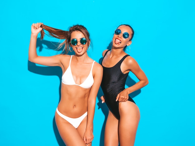Portrait of two beautiful sexy smiling women in summer white and black swimwear bathing suits. Trendy hot models having fun. Girls isolated on blue.Playing with hair in sunglasses