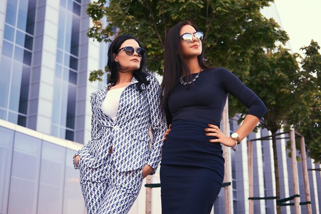 Portrait of two business woman dressed in a stylish formal clothes, standing in a downtown against a background of skyscraper.