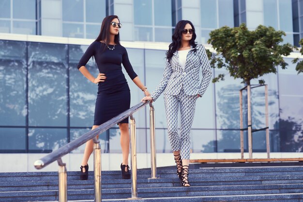 Portrait of two businesswomen dressed in a stylish formal clothes, standing on steps in a downtown posing against a background of a skyscraper.