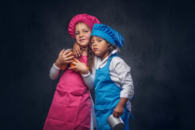 두 귀여운 작은 요리사의 초상화입니다. 파란색 요리복을 입은 갈색 곱슬머리를 한 어린 소년과 분홍색 요리복을 입은 아름다운 여학생이 조리기구와 f를 들고 함께 껴안고 있습니다.