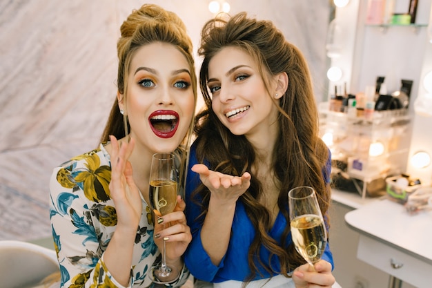 Portrait two fashionable excited happy young women having fun, drinking champagne in haidresser salon