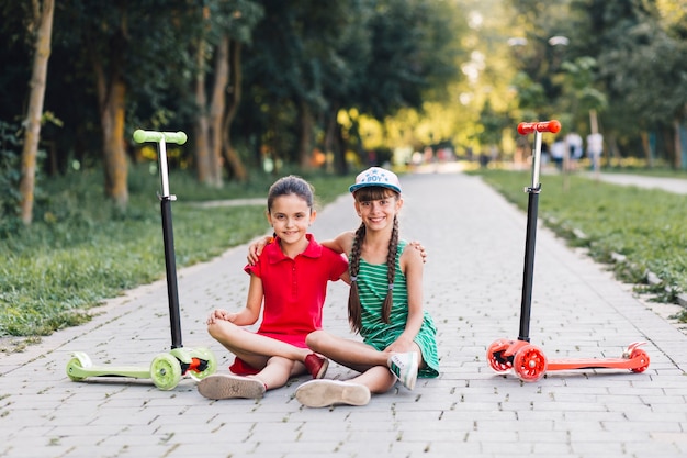 Portrait of two female friends sitting on walkway with their kick scooters in the park