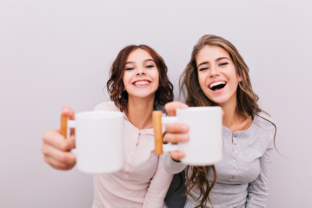 Portrait two funny girls in pajamas having fun on gray wall . They stretching white cups and smiling .