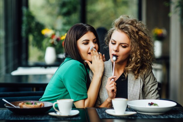 Portrait of two girl friends spend time together drinking coffee in the cafe, having fun by eating dessert, cakes. Said secret for other.