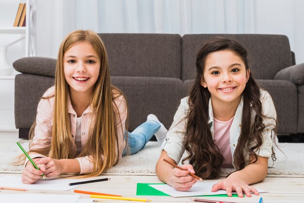 Portrait of two girls laying on carpet drawing color with pencil looking to camera