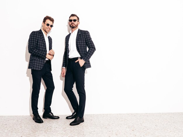 Portrait of two handsome confident stylish hipster lambersexual models Sexy modern men dressed in black elegant suit Fashion male posing in studio near white wall in sunglasses Full length