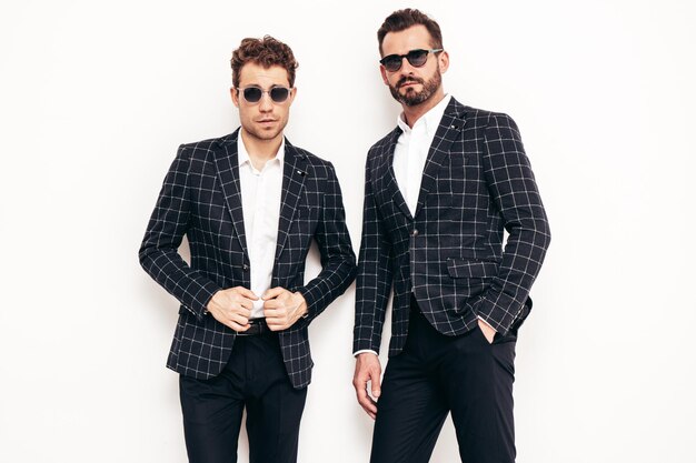 Portrait of two handsome confident stylish hipster lambersexual models Sexy modern men dressed in black elegant suit Fashion male posing in studio near white wall in sunglasses