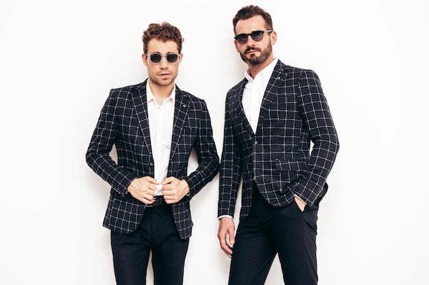 Portrait of two handsome confident stylish hipster lambersexual models Sexy modern men dressed in black elegant suit Fashion male posing in studio near white wall in sunglasses