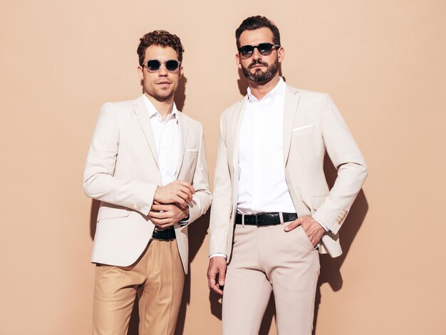 Portrait of two handsome confident stylish hipster lambersexual models Sexy modern men dressed in white elegant suit Fashion male posing in studio near beige wall