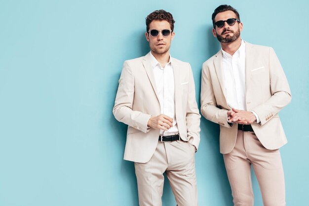 Portrait of two handsome confident stylish hipster lambersexual models Sexy modern men dressed in white elegant suit Fashion male posing in studio near blue wall