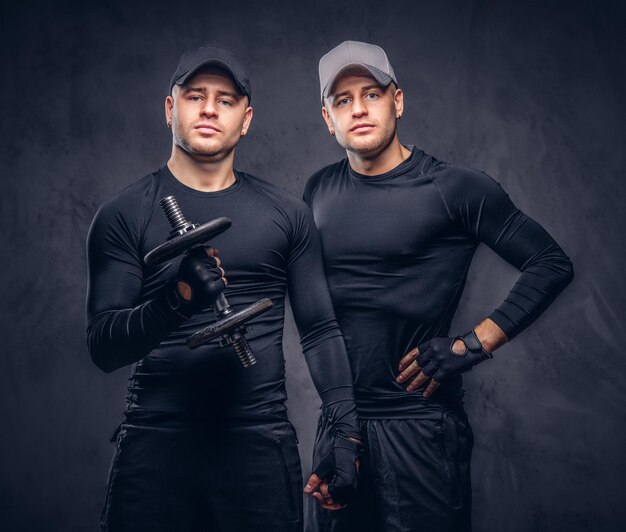 Portrait of two handsome young male dressed in a black sportswear and baseball cap over a dark background, holds a dumbbell.