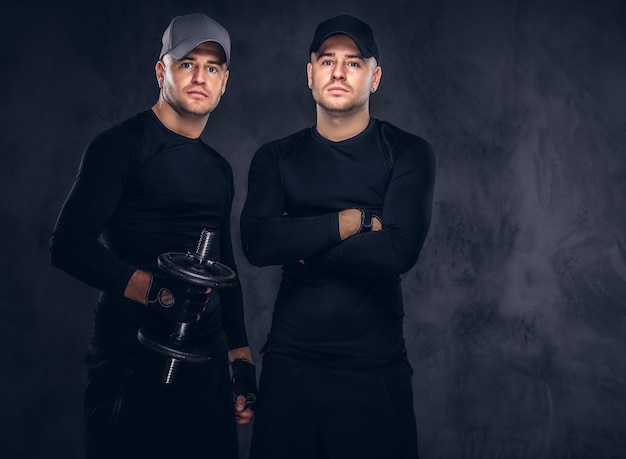 Portrait of two handsome young male dressed in a black sportswear and baseball cap over a dark background, holds a dumbbell.