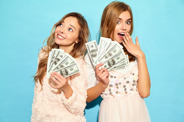 Portrait of two happy delighted blond women wearing summer clothes rejoicing win and holding cash money isolated over blue wall