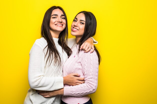 Portrait of two happy girls dressed in sweaters hugging isolated over yellow wall