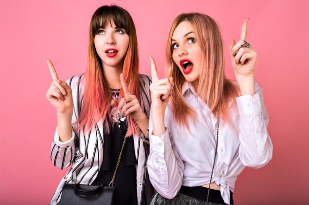  portrait of two happy hipster stylish women showing up by their fingers, exited surprised emotions, pink wall, elegant clothes