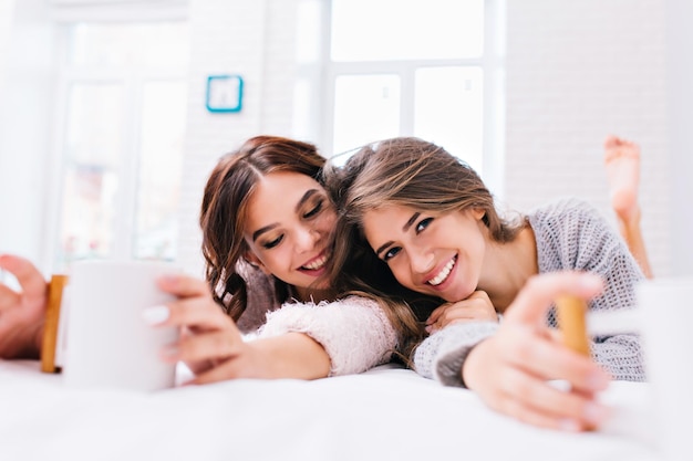 Portrait two joyful smiling young women chilling on bed with cups of tea. waking up in the morning in modern apartment, happy time, good mood