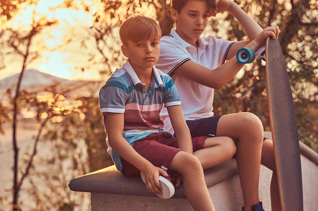 Portrait of two little brothers with a skateboard sitting on the stone guardrail outdoors at a sunset.