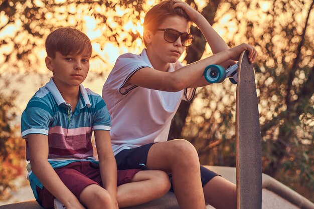 Portrait of two little brothers with a skateboard sitting on the stone guardrail outdoors at a sunset.