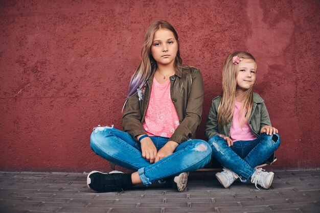 다리 보도에서 스케이트보드에 앉아 있는 동안 벽에 기대어 유행 옷을 입은 두 명의 작은 자매의 초상화.